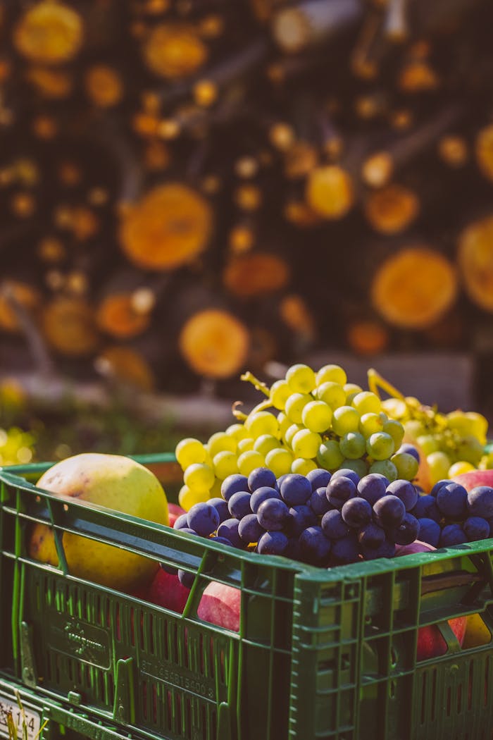 A green crate filled with fresh grapes and pears outdoors with blurred logs in the background.